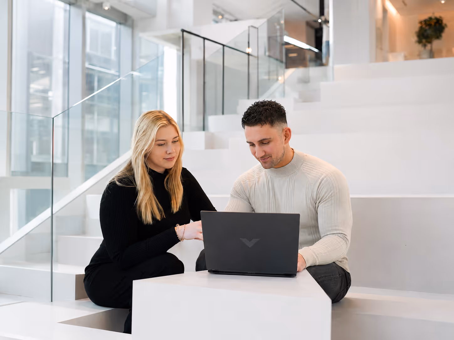 Two people sitting on white steps working together on a laptop in a modern bright space.