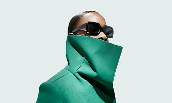 Woman in oversized green coat and large black sunglasses against a light background.
