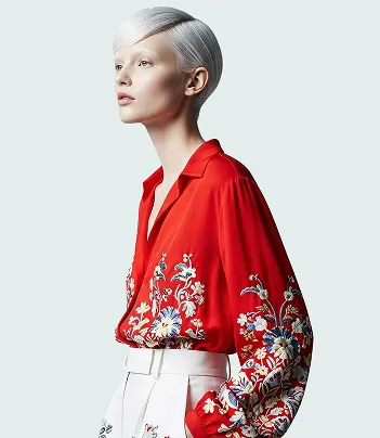 Model with short silver hair wearing a red blouse with floral embroidery and white pants.
