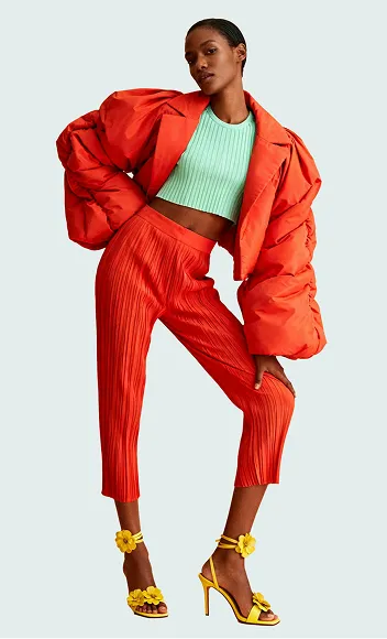 Model wearing bright orange pants and puffy jacket with mint green cropped top and yellow heels with flowers.