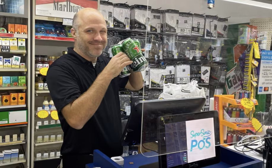 Smiling convenience store clerk holding beer cans at a SuperSonic POS counter