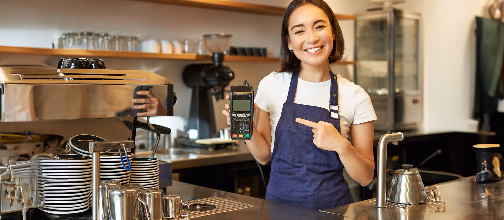 Smiling barista in an apron holding and pointing at a card payment terminal in a cafe