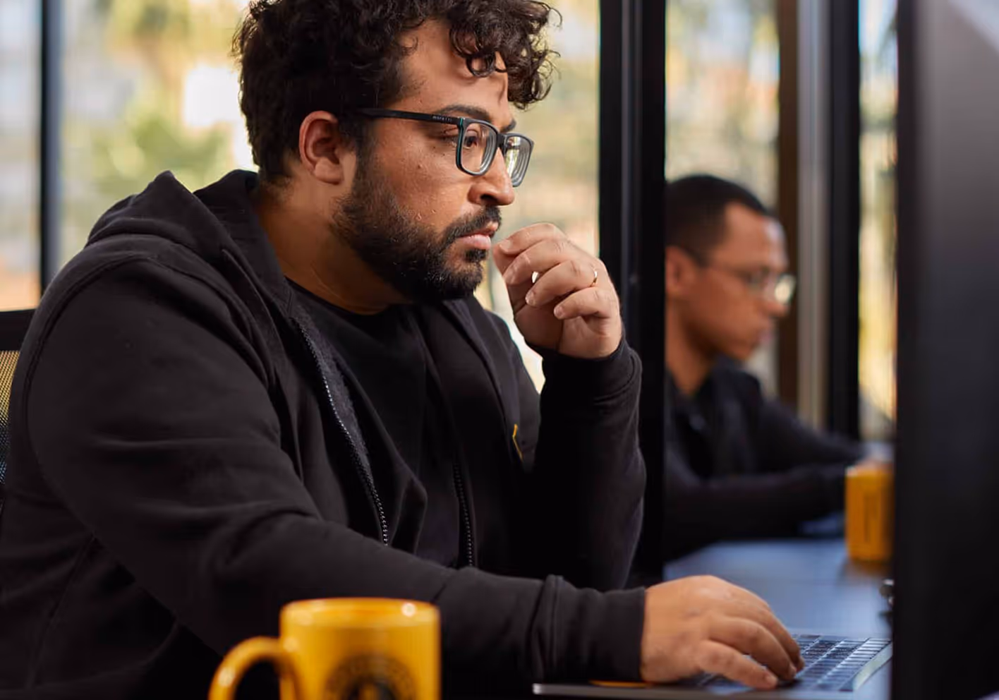 Man with glasses and beard focused on laptop typing, with blurred person working in background.