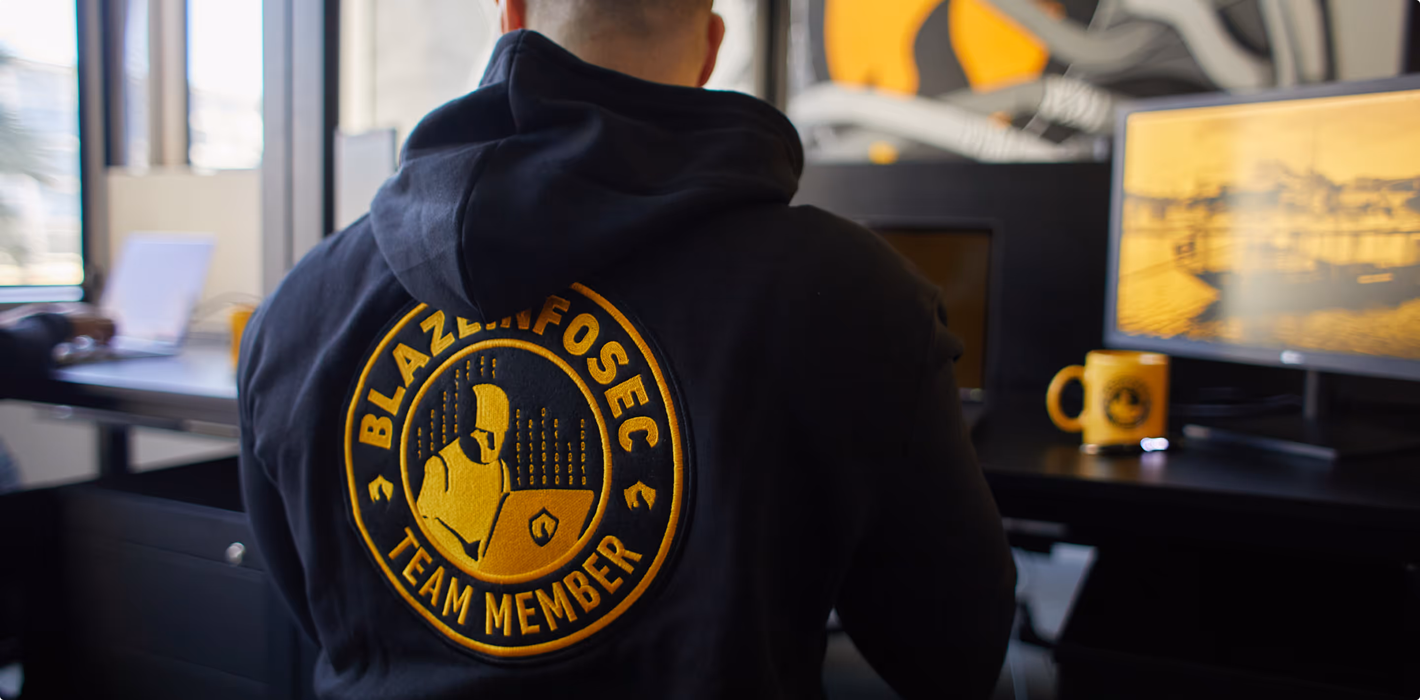 Person wearing a black hoodie with BlazeInfoSec team member logo sitting at a desk with a monitor and mug.