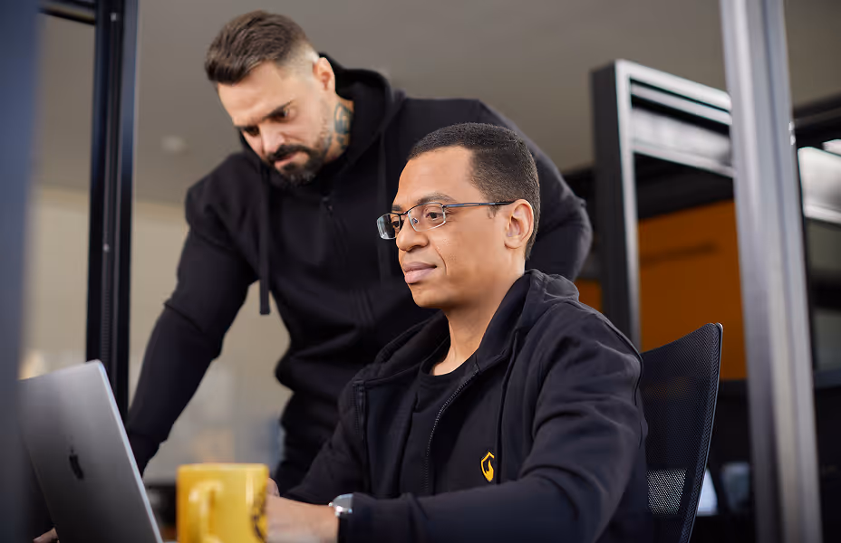 Two men in black hoodies working together looking at a laptop screen in an office.
