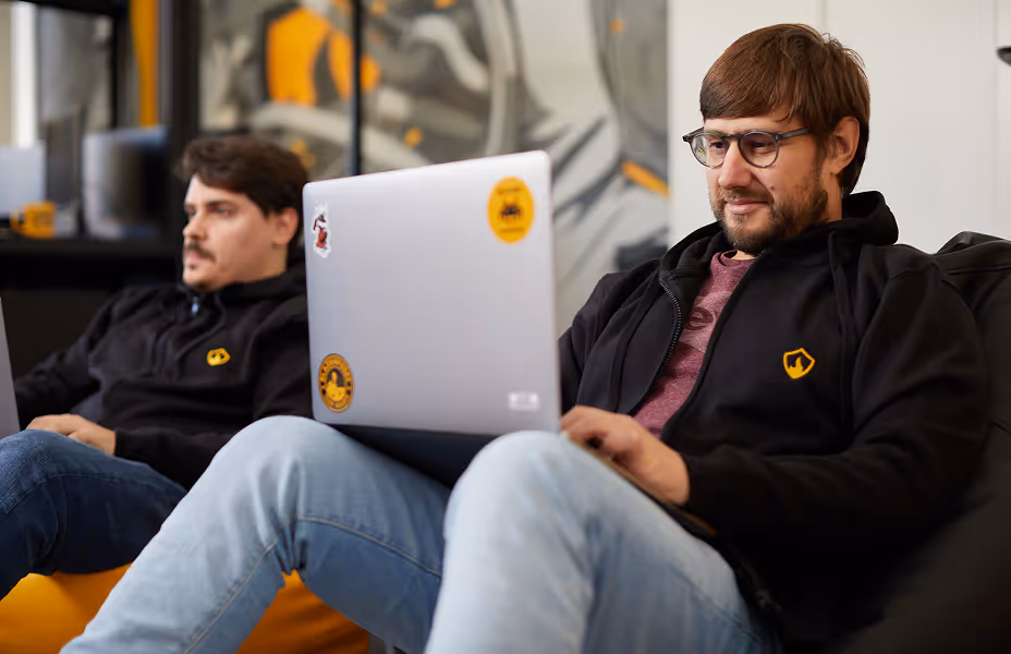 Two men in black hoodies sitting indoors, one focused on a laptop with stickers in a relaxed setting.