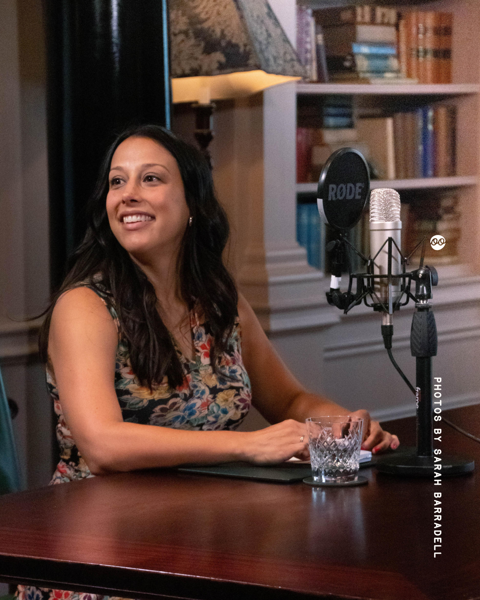 A smiling woman with long dark hair wearing a sleeveless floral dress, sitting at a wooden table with a glass of water and a microphone on a stand in front of her, against a background of bookshelves and a lamp.