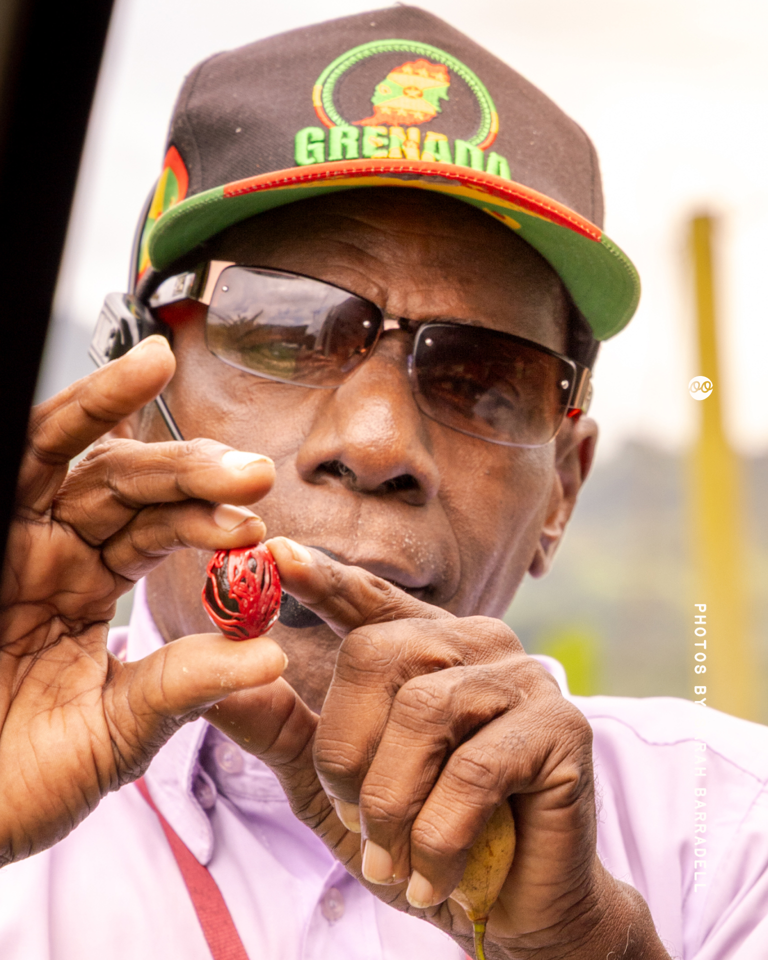 Close-up of a man wearing sunglasses and a black cap with Grenada logo holding a red and black nutmeg seed with his fingers while showing it to the camera.