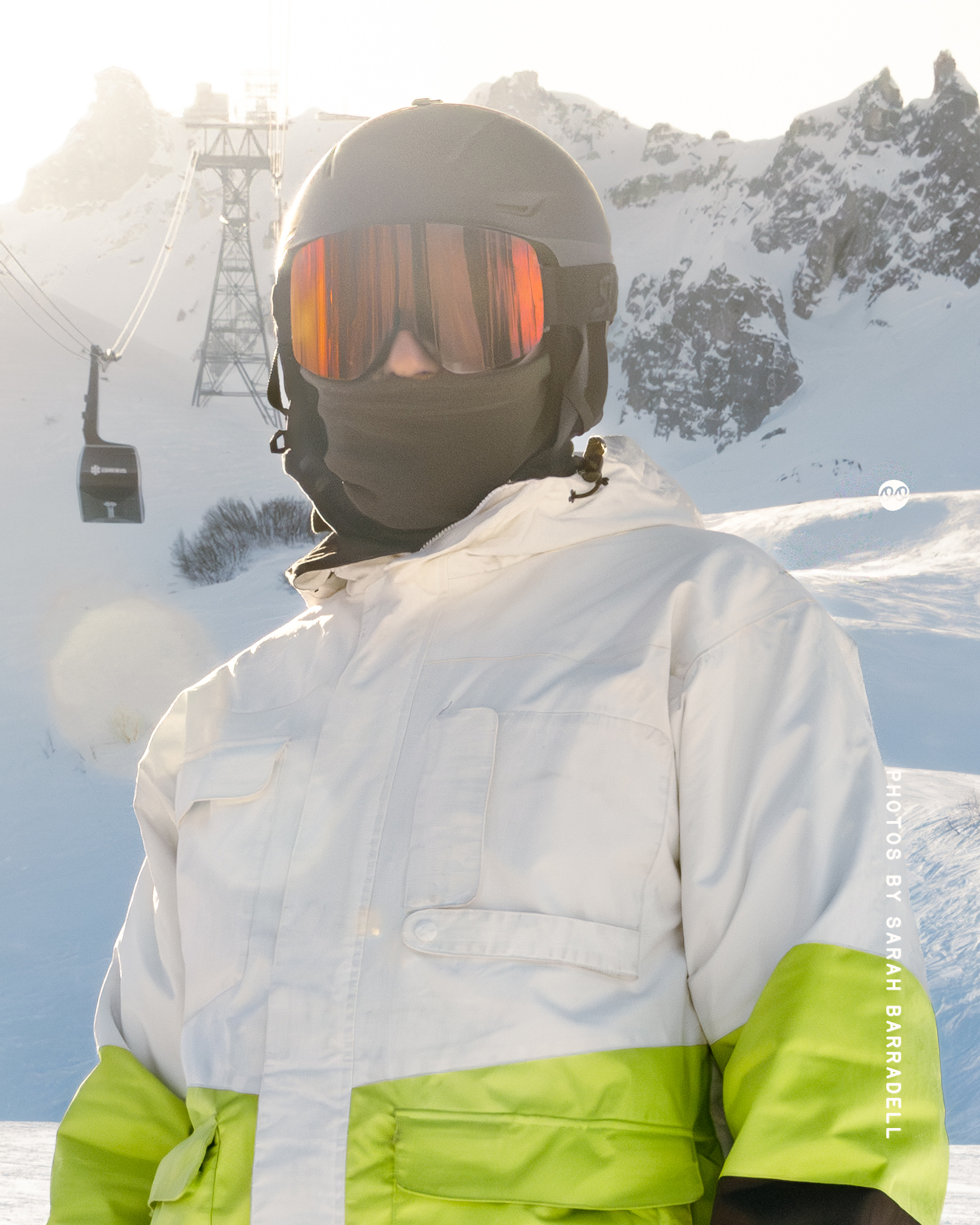 Person in white and green ski jacket wearing black helmet and reflective orange ski goggles standing in snowy mountain landscape with ski lift in the background.