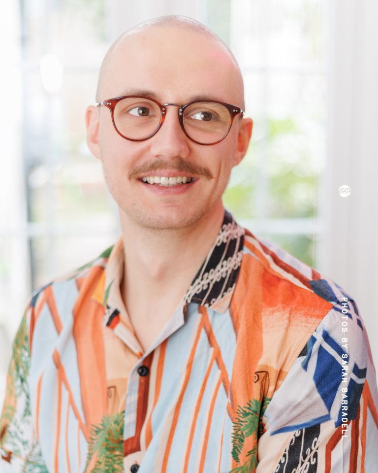 Smiling man with a mustache and glasses wearing a colorful patterned shirt with orange, blue, and green tones, standing in a bright indoor setting with blurred background.