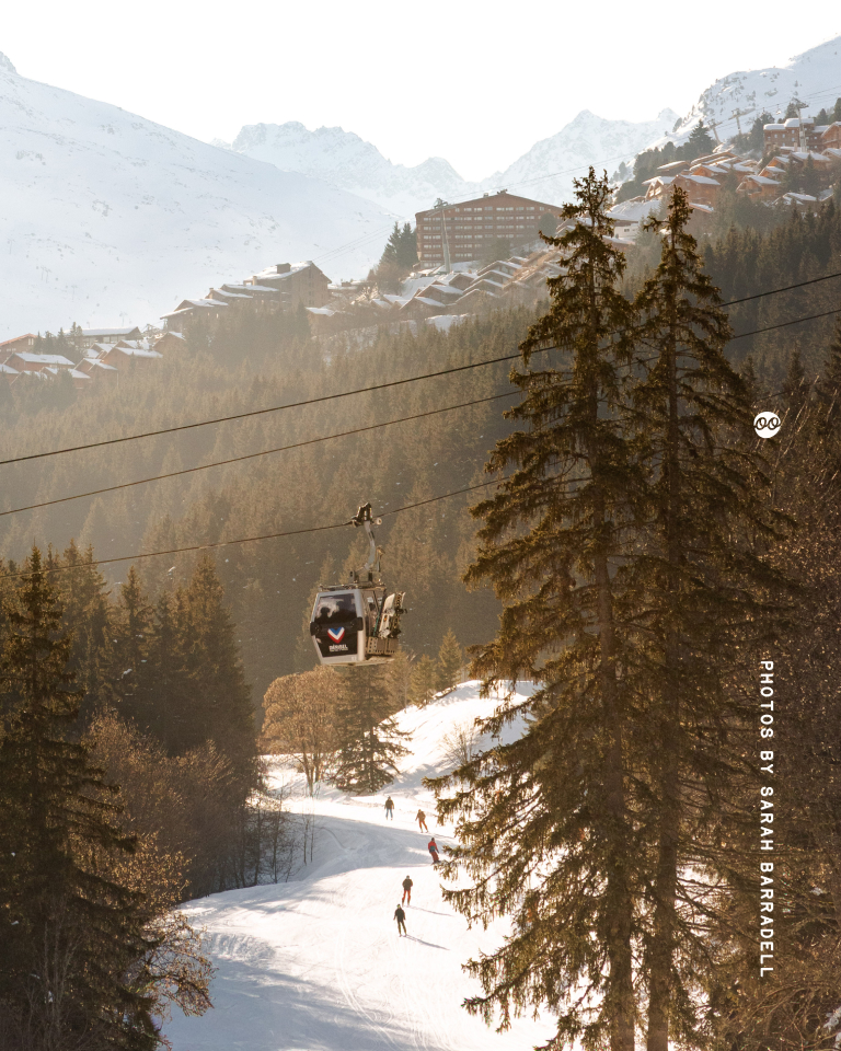 Skiers descend a snow-covered slope surrounded by tall pine trees with a gondola cable car above and snow-capped mountains and buildings in the background bathed in soft sunlight.