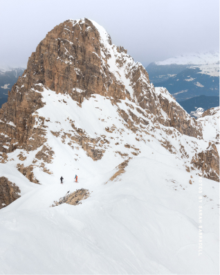 Two hikers walking on a snowy mountain slope below a rugged, rocky peak under a cloudy sky.