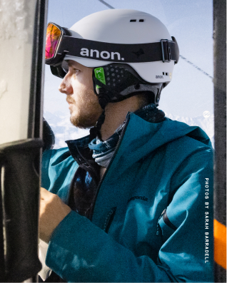 Profile view of a man wearing a white helmet with black 'anon.' goggles and a blue ski jacket, looking out of a window.