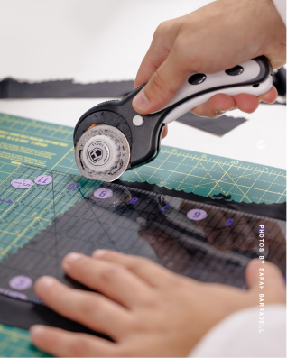 Close-up of hands using a rotary cutter to slice black fabric on a green cutting mat with measurement grid and a transparent ruler. From a shoot with London-based Demeter Bio, who are creating a bio-based PFAS alternative.