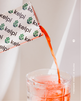 A container labeled 'kelpi' pouring a bright orange liquid into a clear glass filled with ice. From a shoot with Kelpi, who are creating a seaweed-based alternative to plastic sachets. 