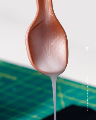 Close-up of a brown plastic spoon dripping with white liquid over a green cutting mat with grid lines. From a shoot with London-based Demeter Bio, who are creating a bio-based PFAS alternative.