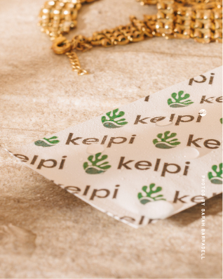 Close-up of a white fabric with a repeating green hand logo and the word 'Kelpi' printed on it, lying on a light brown surface with a gold chain in the background. From a shoot with Kelpi, who are creating a seaweed-based alternative to plastic sachets. 
