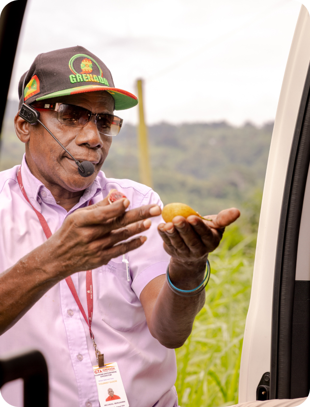 Man wearing a black cap with Grenada logo, sunglasses, and a headset microphone, holding two small fruits in his hands, standing near a vehicle with a blurred green landscape in the background.
