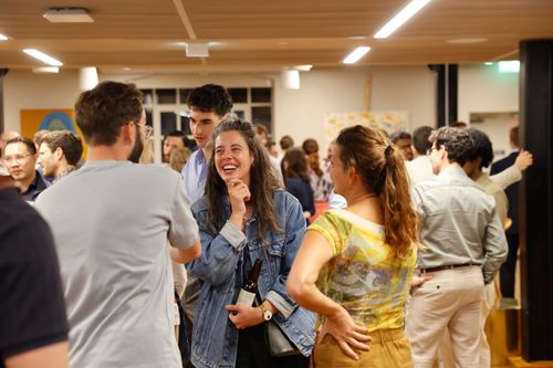 A group of people socializing indoors, focusing on a smiling Sarah Barradell in a denim jacket, engaged in conversation with a man in a gray shirt and another woman in a yellow patterned top.