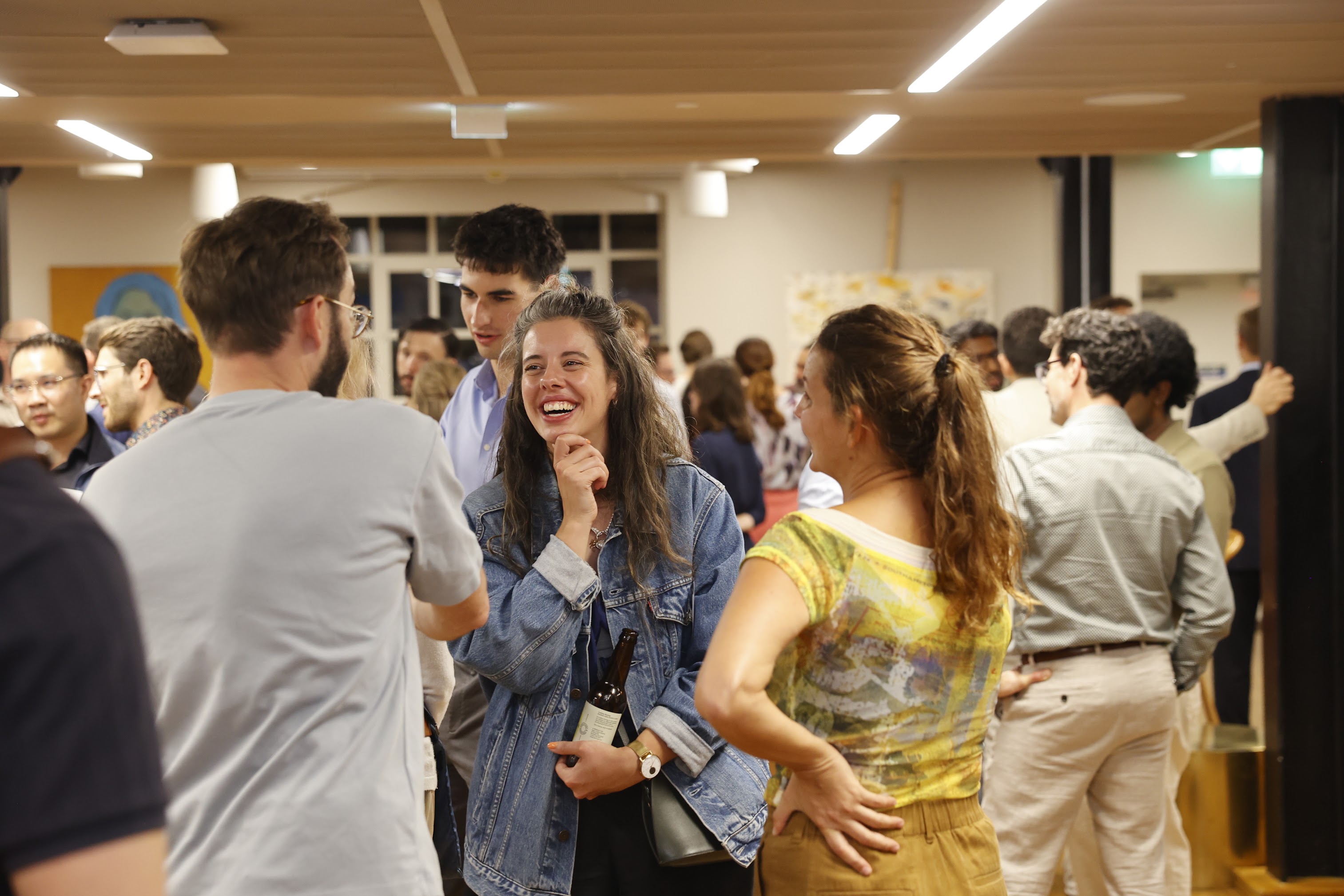 A group of people socializing indoors, focusing on a smiling Sarah Barradell in a denim jacket, engaged in conversation with a man in a gray shirt and another woman in a yellow patterned top.