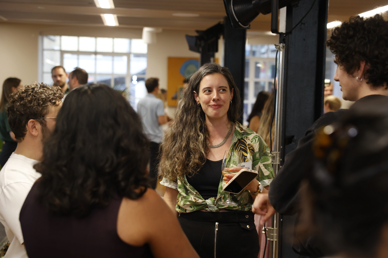 Sarah Barradell with long curly hair wearing a patterned green shirt and black skirt holds a glass of wine and a phone while talking to a group of people in a well-lit room with large windows.