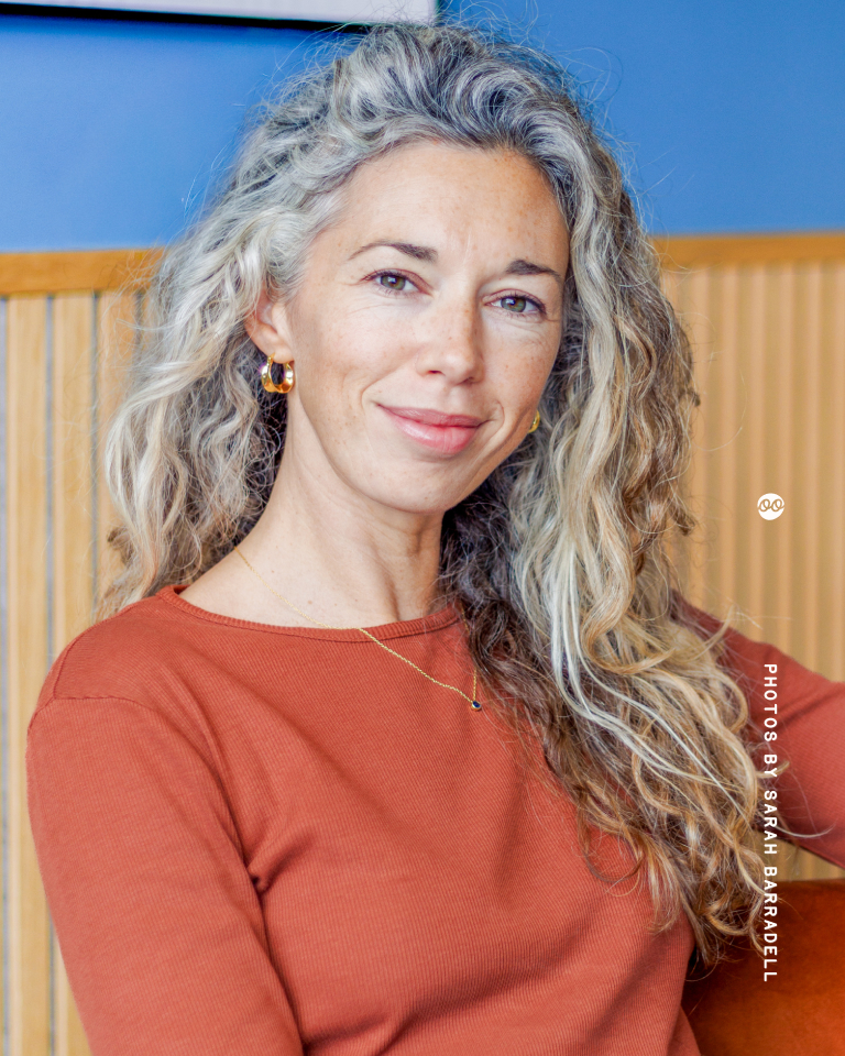 Woman with long, curly gray and blonde hair wearing a burnt orange top, gold hoop earrings, and a delicate necklace, smiling softly against a background of wooden and blue panels.