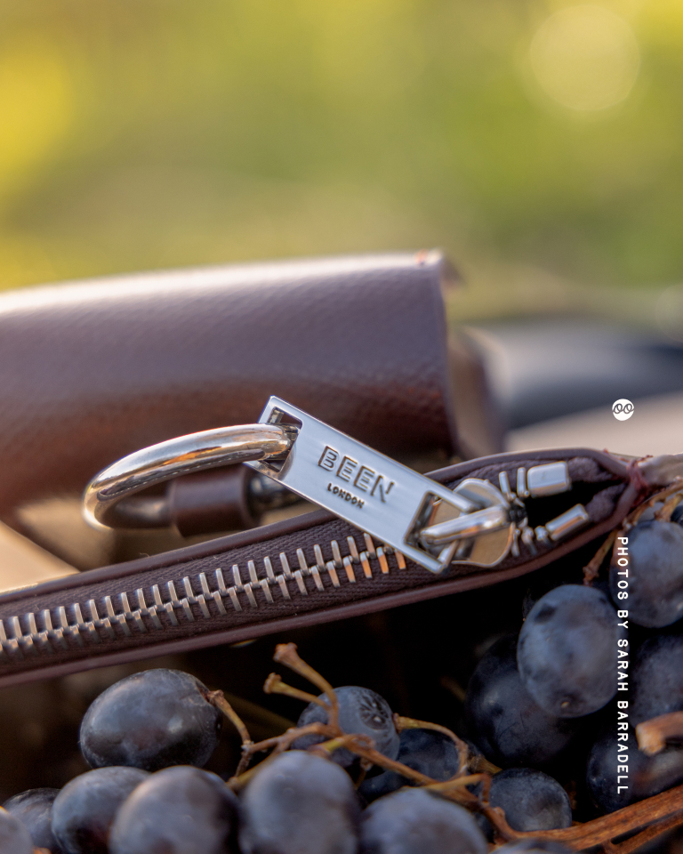 Close-up of a brown leather bag zipper with a silver zipper pull engraved with 'BEEN LONDON,' surrounded by dark purple grapes.