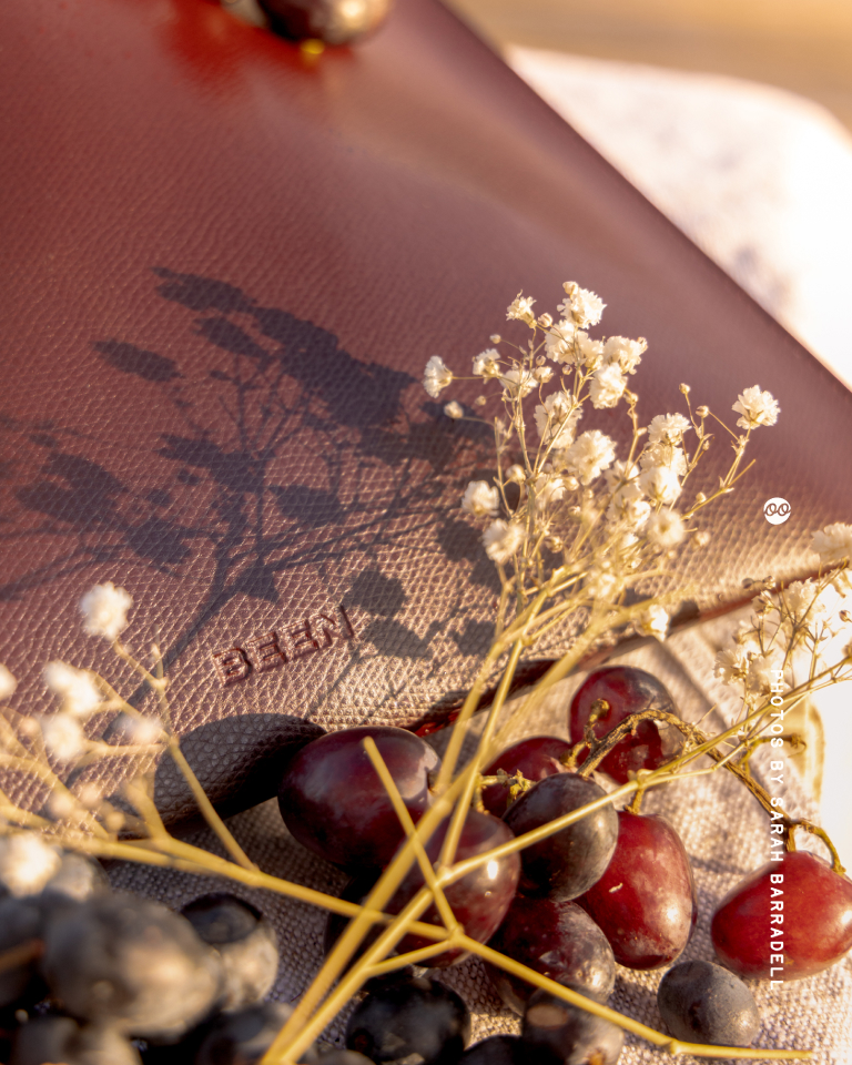 Close-up of a textured brown leather surface with the word 'BEEN' embossed on it, surrounded by small white dried flowers and clusters of dark red and purple grapes casting shadows in warm sunlight.