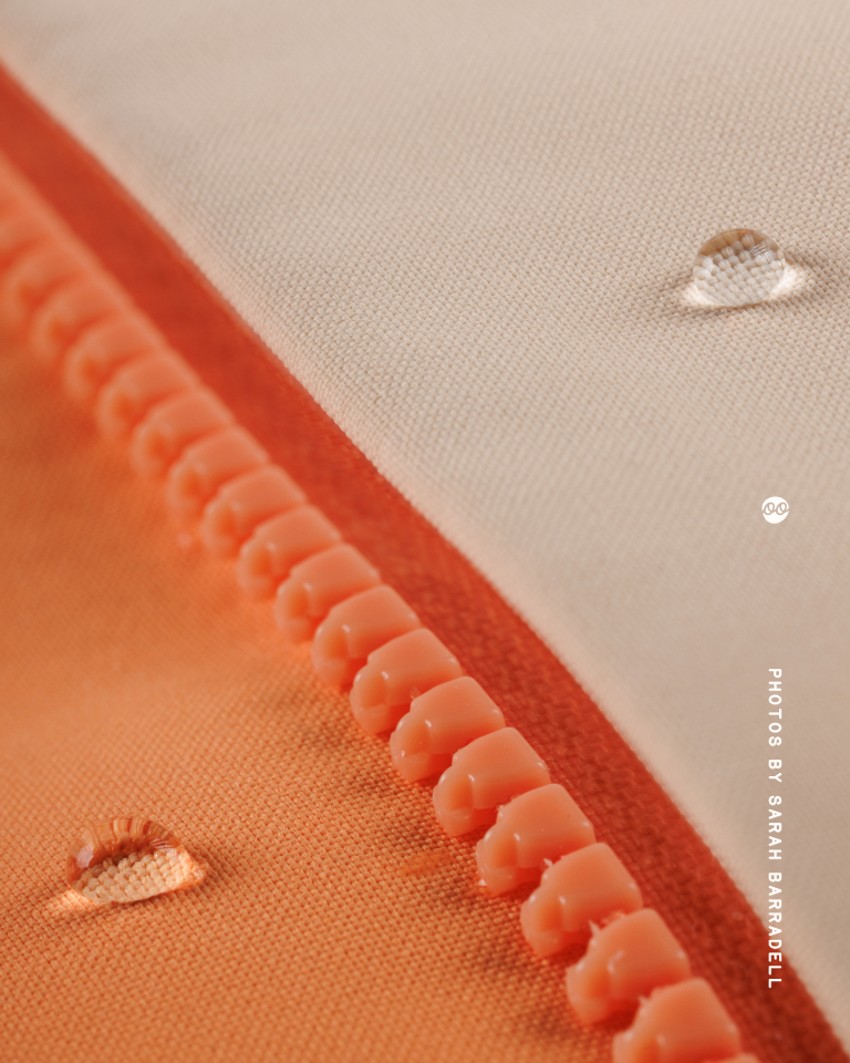 Close-up of a coral orange zipper on orange fabric with water droplets on both orange and cream-colored fabric sections.