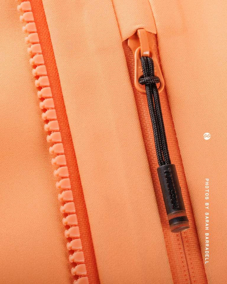 Close-up of an orange zipper with a black pull cord attached to the slider, showing the texture of the fabric and teeth of the zipper.