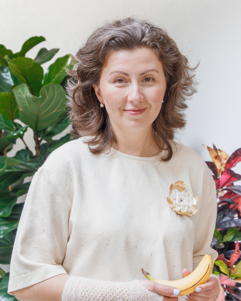 A woman with shoulder-length curly brown hair wearing a light beige sweater with a decorative flower pin, holding a banana, standing indoors with green and red leafy plants in the background.