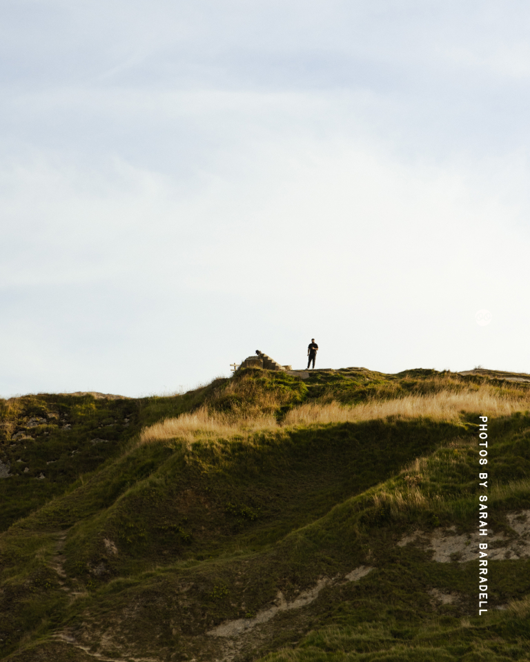 A solitary person standing on top of a grassy hill under a pale sky, viewed from below.