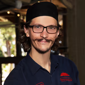 Smiling man with glasses and a mustache wearing a black hat and navy work shirt indoors.