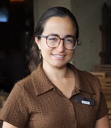 Woman with glasses and long dark hair wearing a brown textured shirt and name tag reading RACHEL.