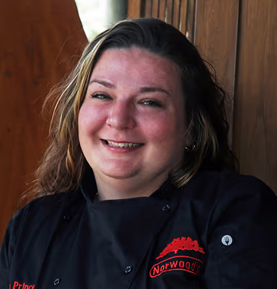 Smiling woman with shoulder-length hair wearing a black Norwoods chef jacket against a wooden background.