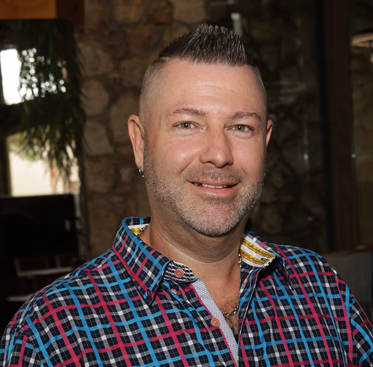 Smiling man with short spiked hair and a trimmed beard wearing a colorful plaid shirt indoors.