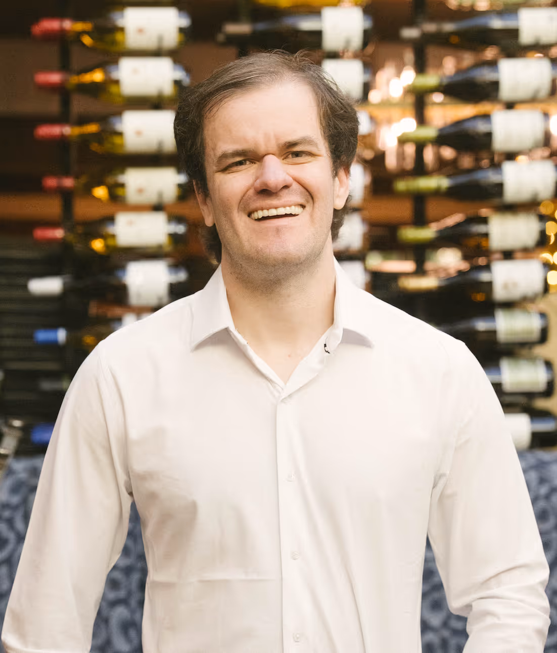 Smiling man in a white shirt standing in front of a wine bottle wall display.
