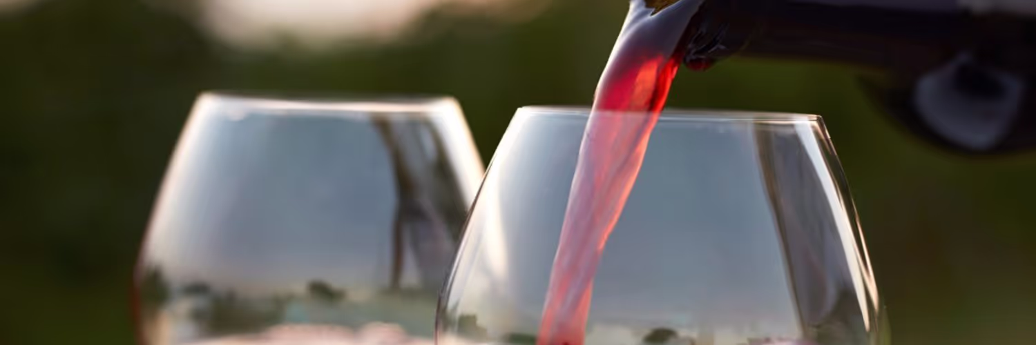 Red wine being poured into a glass with another glass blurred in the background outdoors.