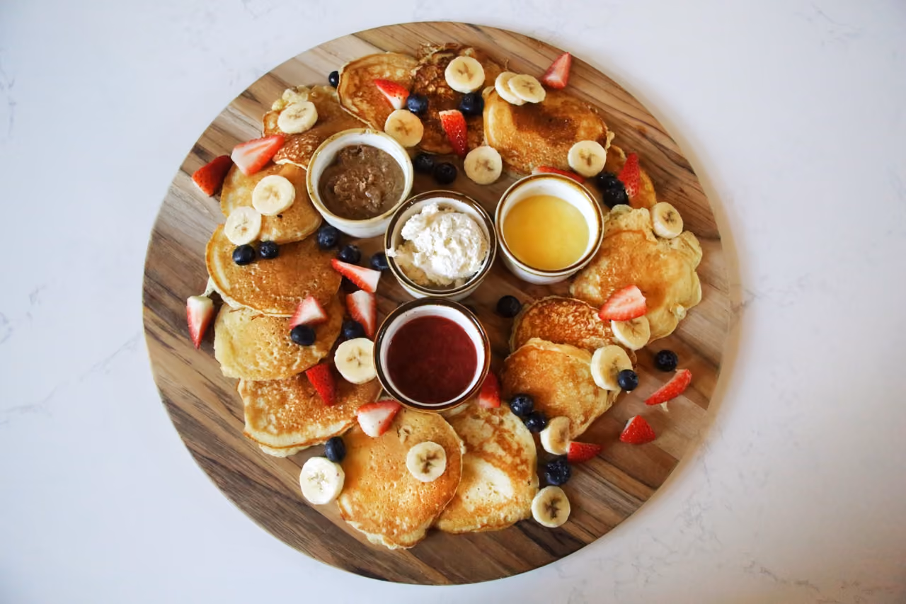 Wooden board with pancakes topped with banana, strawberry, blueberry, and three small bowls of sauces.