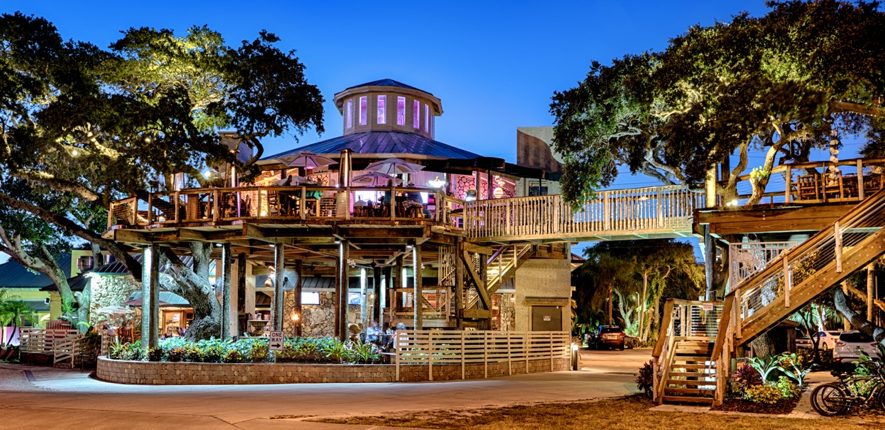Night view of a two-level wooden restaurant with outdoor seating surrounded by trees and lights.