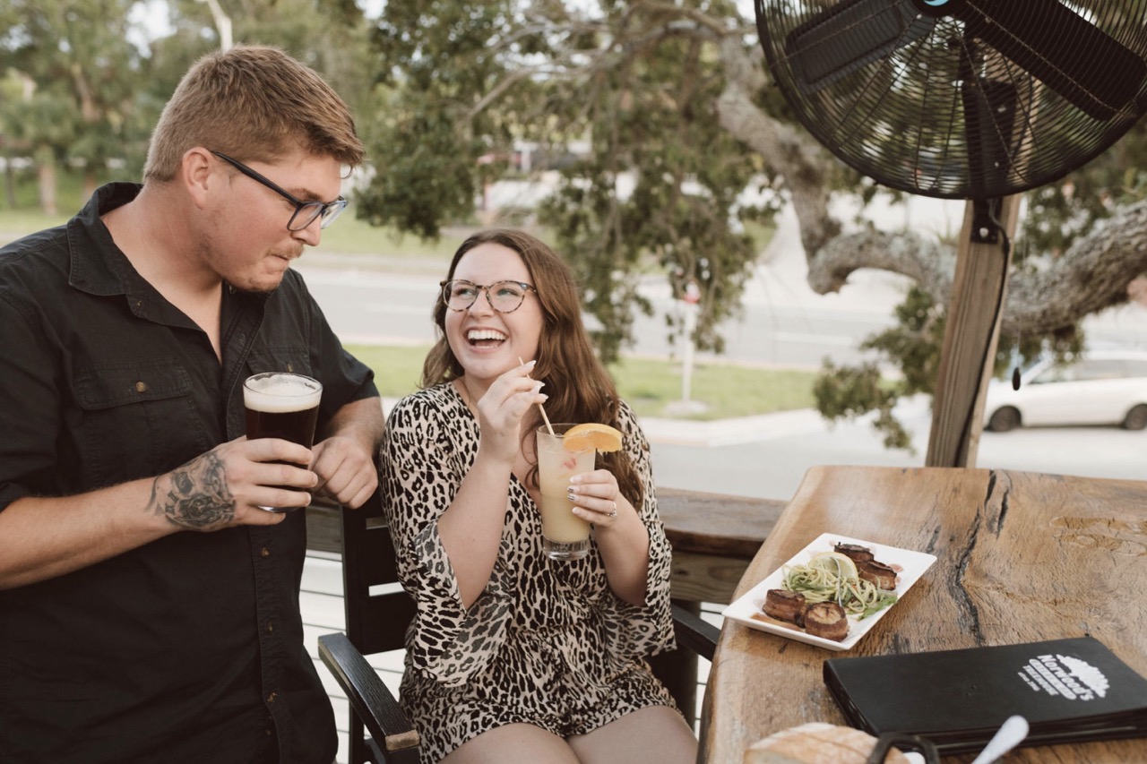 Couple laughing and holding drinks on an outdoor patio with food on the table.