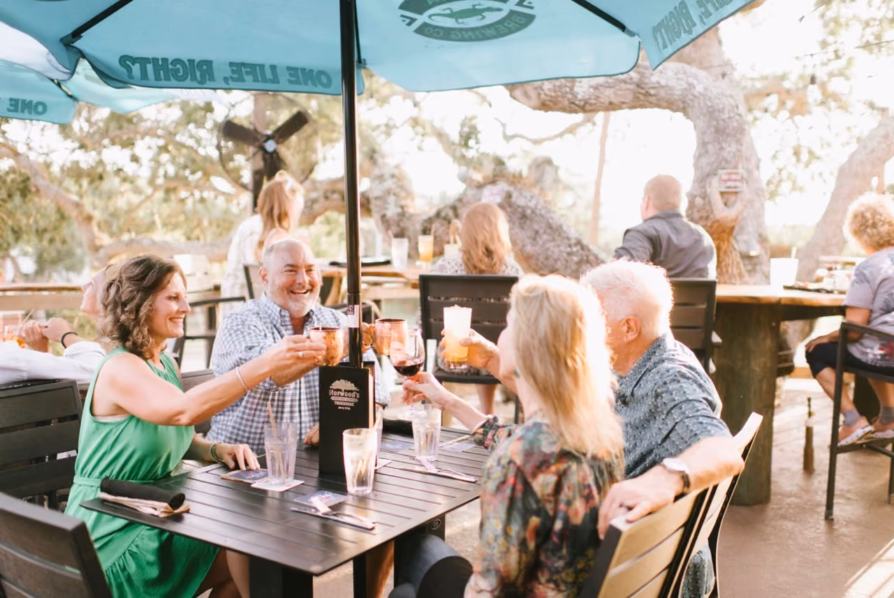 Group of four adults toasting drinks together at an outdoor patio under umbrellas.