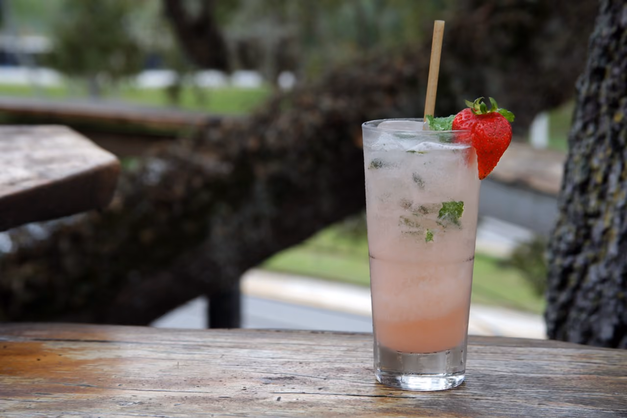 Tall glass of pink mojito with ice, mint leaves, and a strawberry garnish on a wooden table outdoors.