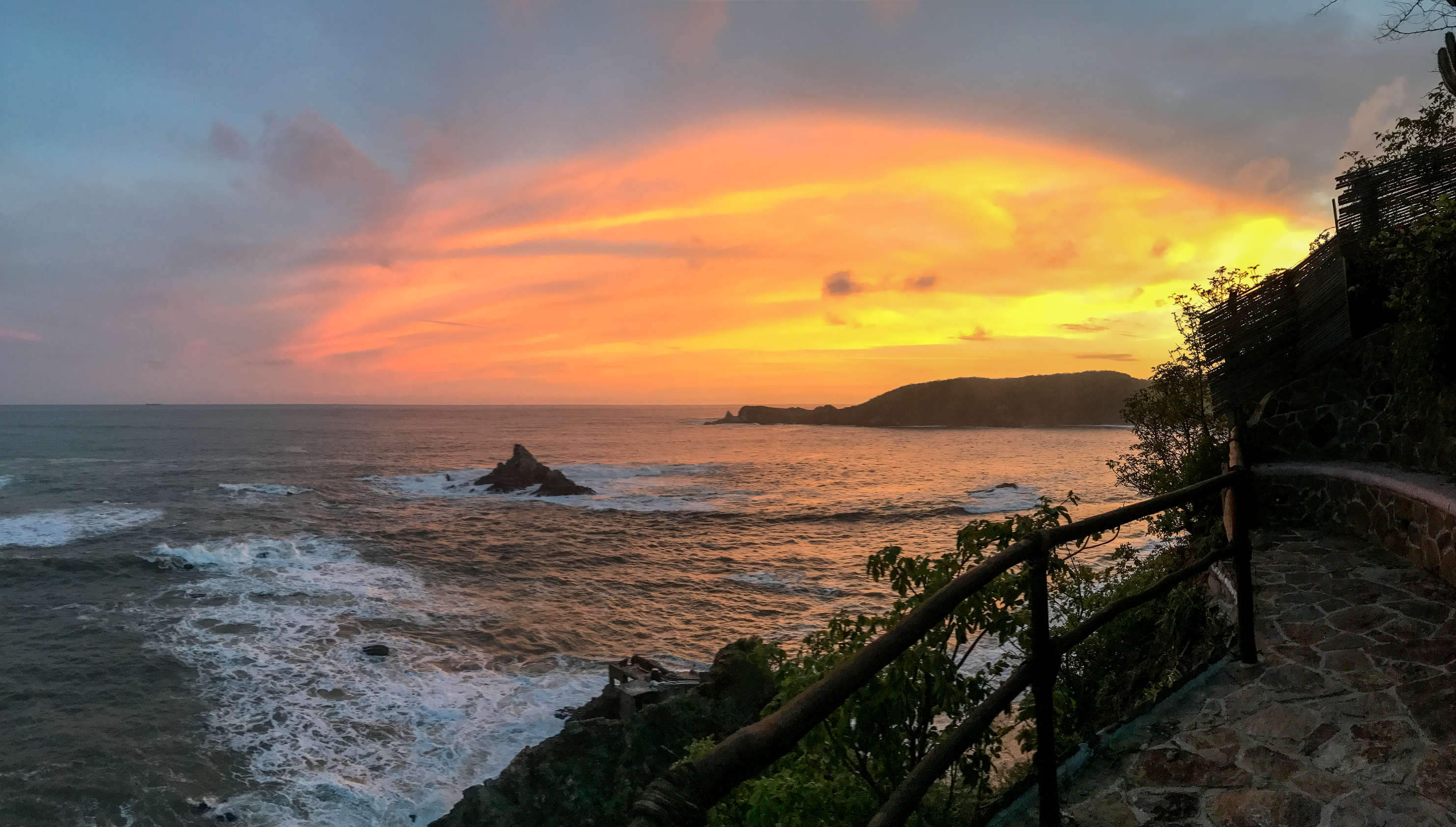 Atardecer sobre las olas del océano con costa rocosa y un sendero de piedra con barandilla a la derecha.
