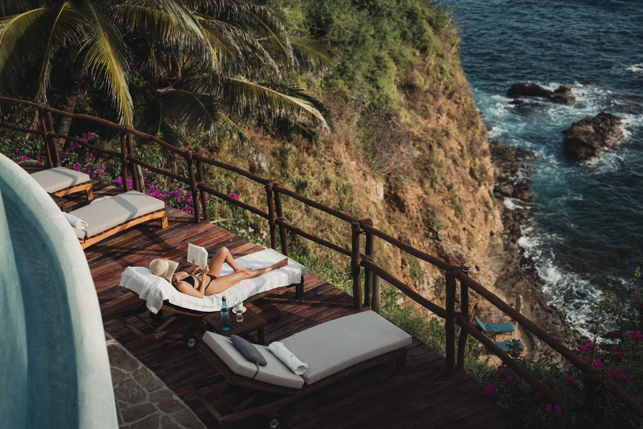 Mujer con sombrero leyendo en una tumbona junto a una piscina con vistas a acantilados rocosos y olas del océano.