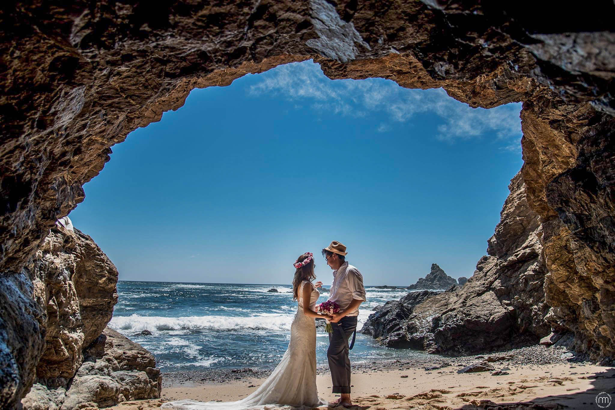 Novios tomados de la mano en una playa, enmarcados por la entrada de una cueva rocosa con olas del océano de fondo.
