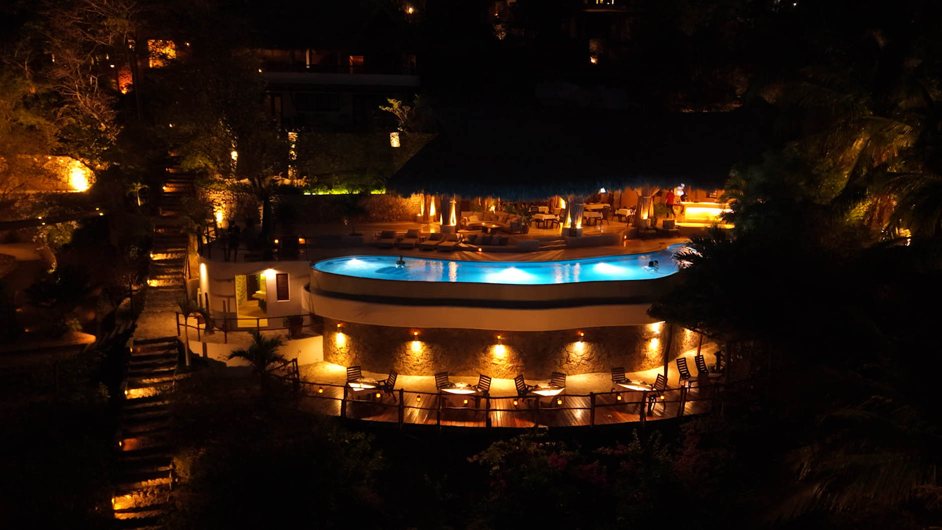 Night view of a lit outdoor pool with people swimming and seating area with tables and chairs below.