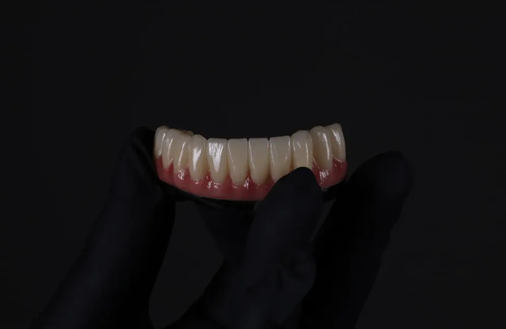 Hand in black glove holding a dental prosthesis with teeth and pink gums against a black background.
