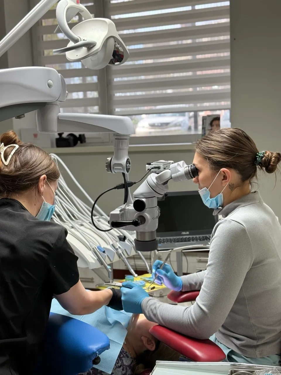 Two masked dental professionals working on a patient's mouth using a dental microscope and tools.