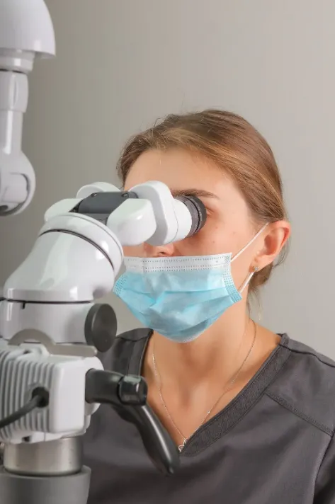 Woman wearing a mask looking through a microscope in a clinical or laboratory setting.